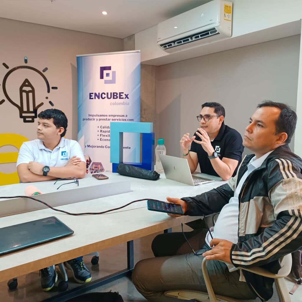 Tres personas sentadas alrededor de una mesa de conferencias en una oficina moderna, con portátiles y materiales de trabajo, frente a un banner de ENCUBEx Colombia que promueve calidad, rapidez, flexibilidad y economía, acompañado de un gráfico de bombilla con lápiz.